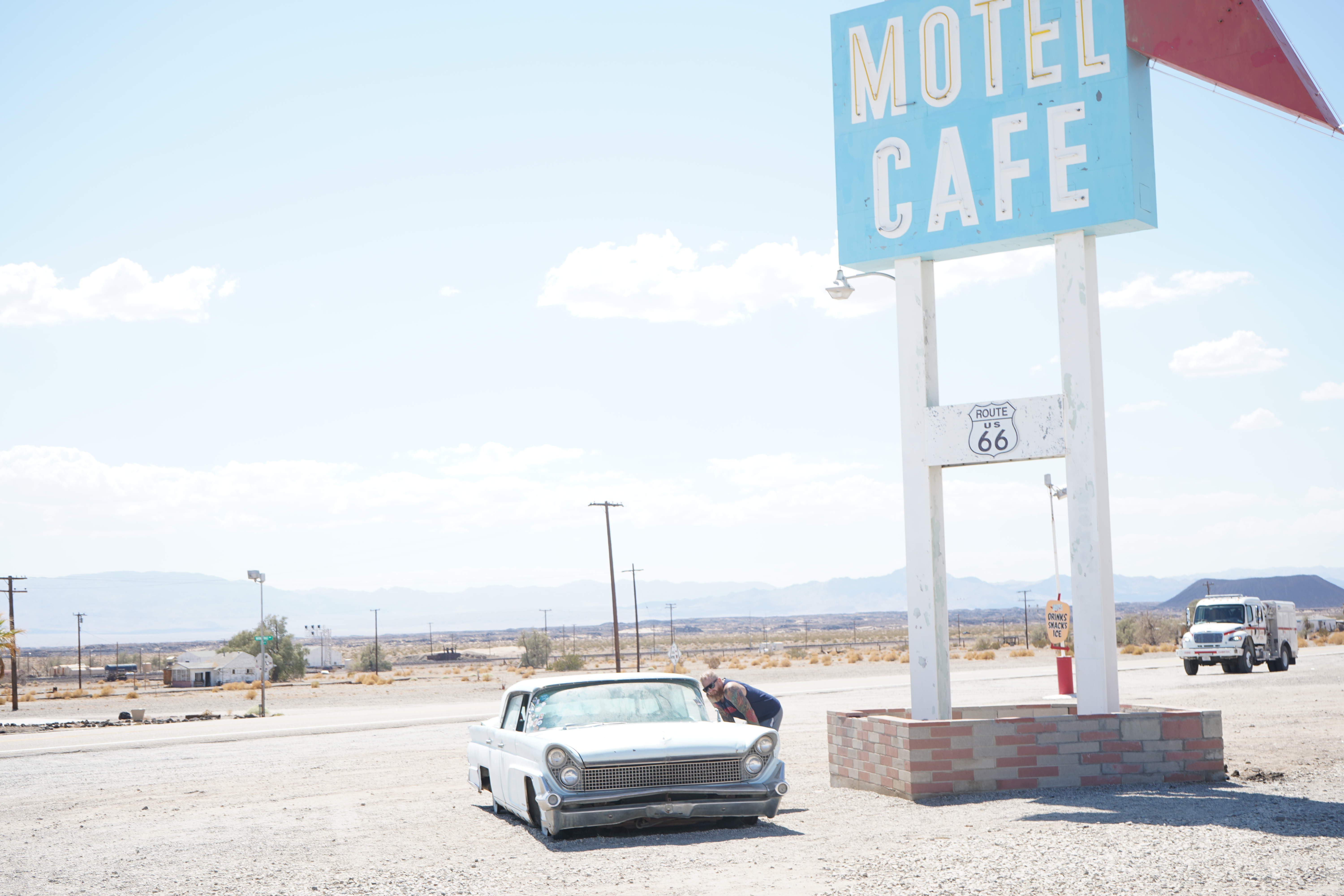 Chris Martin leaning into a classic Lincoln Continental at the Route 66 Motel Cafe sign — Mojave Desert